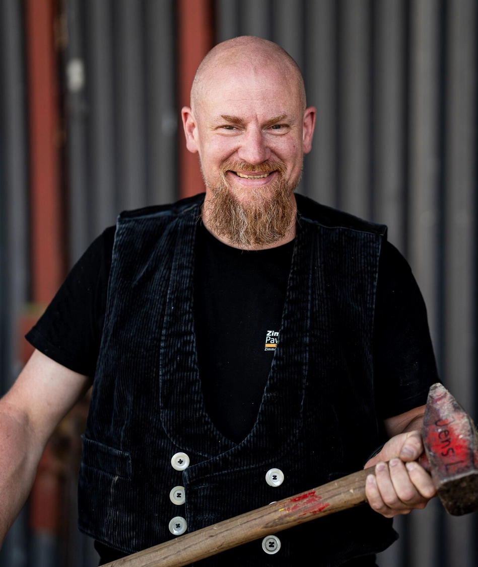 Alexander Feld, ein lächelnder Zimmermann in traditioneller Arbeitskleidung, hält einen Hammer und blickt freundlich in die Kamera.