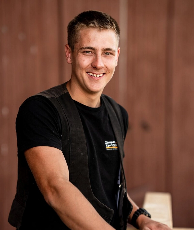 Junger Zimmermann Benjamin Glindmeier lächelt freundlich in die Kamera, im Hintergrund eine Holzfassade.