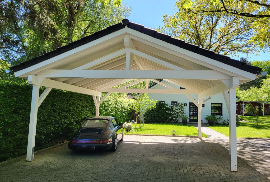 Weißer Holzcarport mit dunklem Dach über einem klassischen Porsche auf einer gepflasterten Einfahrt, umgeben von Grünflächen und einem Haus.