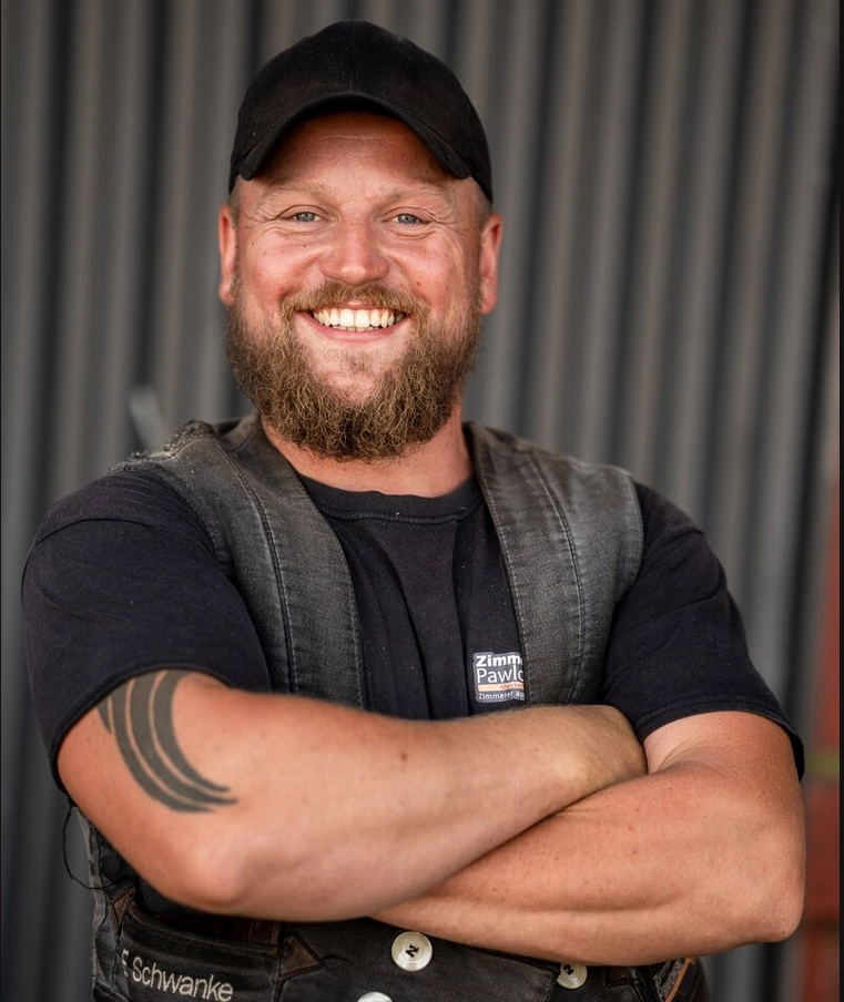 Eric Schwanke, ein lächelnder Zimmermann der Zimmerei Pawlowski GmbH, mit Bart, Mütze und Arbeitskleidung, die Arme verschränkt.