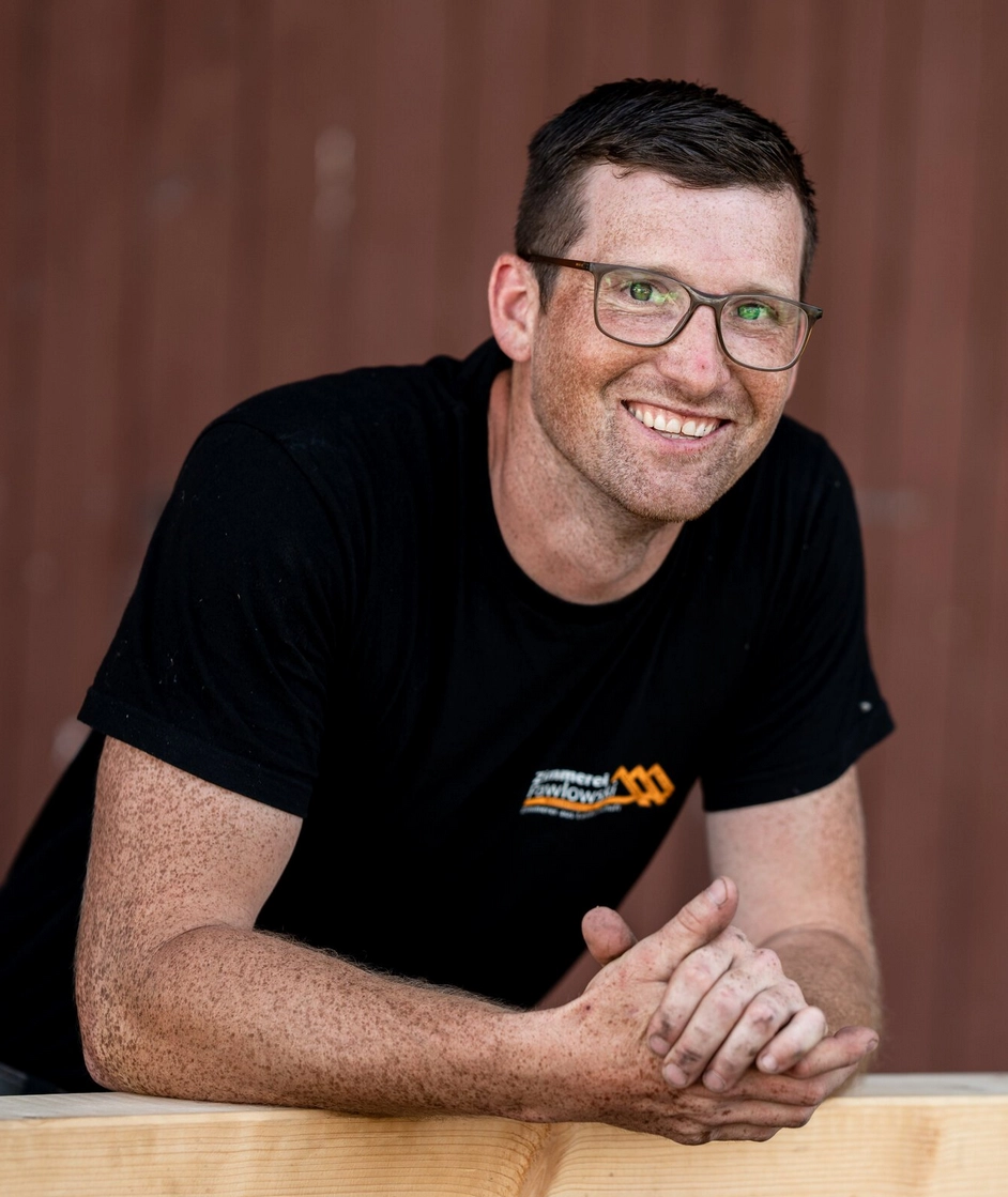 Fabian Steinfeld, ein lächelnder Zimmermann mit Brille, stützt sich in Arbeitskleidung auf einen Holzbalken und blickt freundlich in die Kamera.