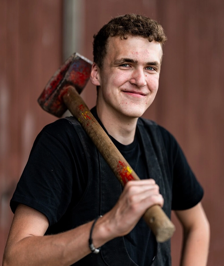 Henrik Jacobsen, ein junger Zimmermann, lächelt freundlich in die Kamera, während er einen großen Hammer auf seiner Schulter trägt.