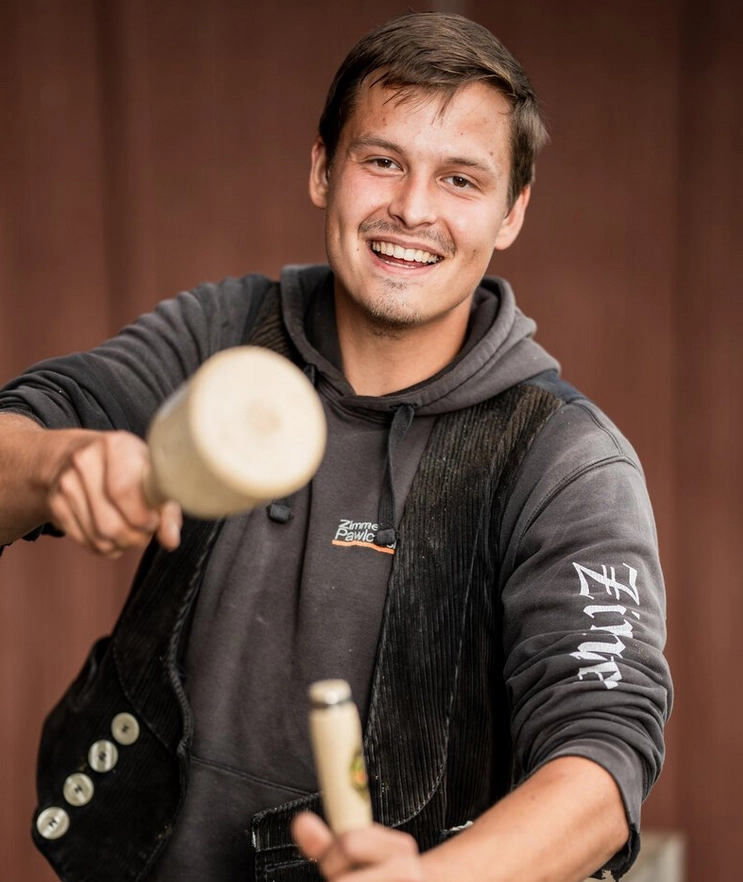 Jermain Erdmann, ein lächelnder Zimmermann in traditioneller Kluft, hält zwei Holzschlegel und repräsentiert das Handwerk der Zimmerei.