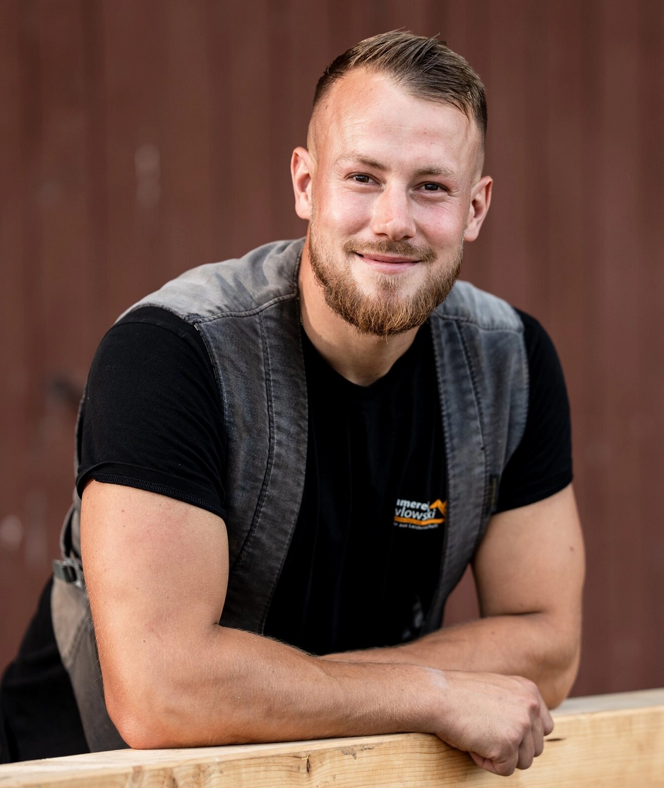 Porträt von Ken Pawlowski, einem lächelnden Mitarbeiter der Zimmerei Pawlowski, der sich in Arbeitskleidung auf einen Holzbalken lehnt.