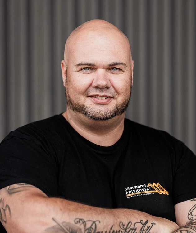 Porträt von Marcel Rammelsberg, einem Mitarbeiter der Zimmerei, der lächelnd in die Kamera blickt und ein T-Shirt mit dem Firmenlogo trägt.