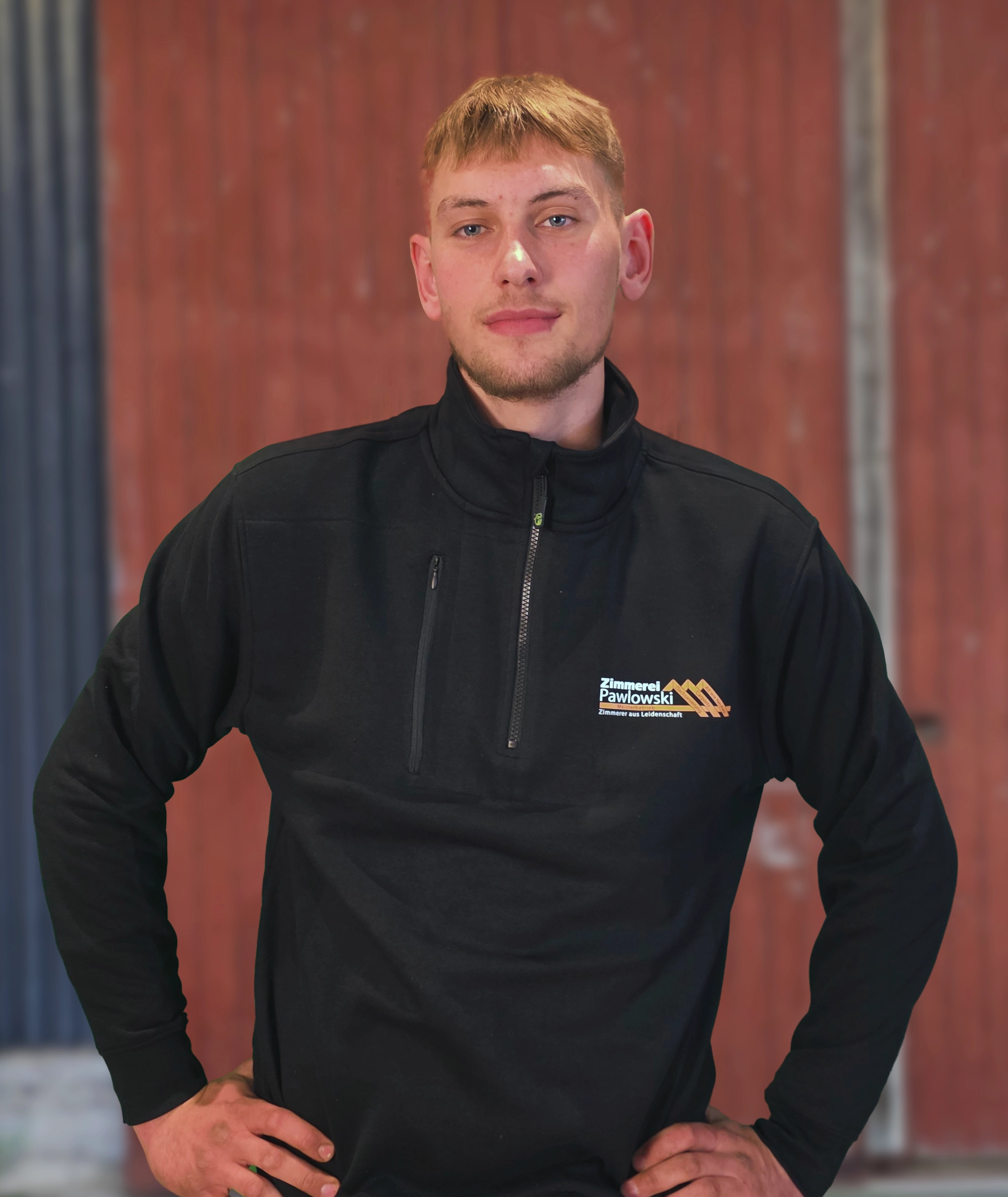 Paul Silaev, Mitarbeiter der Zimmerei Pawlowski GmbH, trägt eine schwarze Arbeitsjacke mit Firmenlogo und steht vor einer roten Wand.