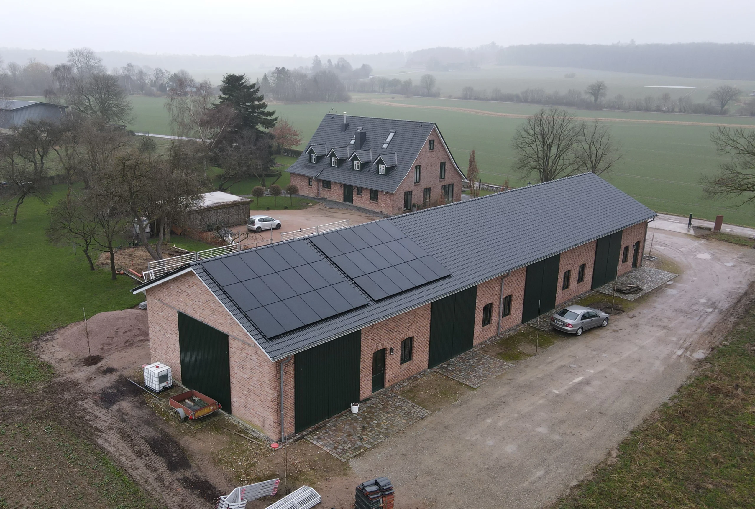 Luftaufnahme eines modernen Wohnhauses und einer Scheune mit Photovoltaikanlage auf dem Dach, umgeben von ländlicher Natur.