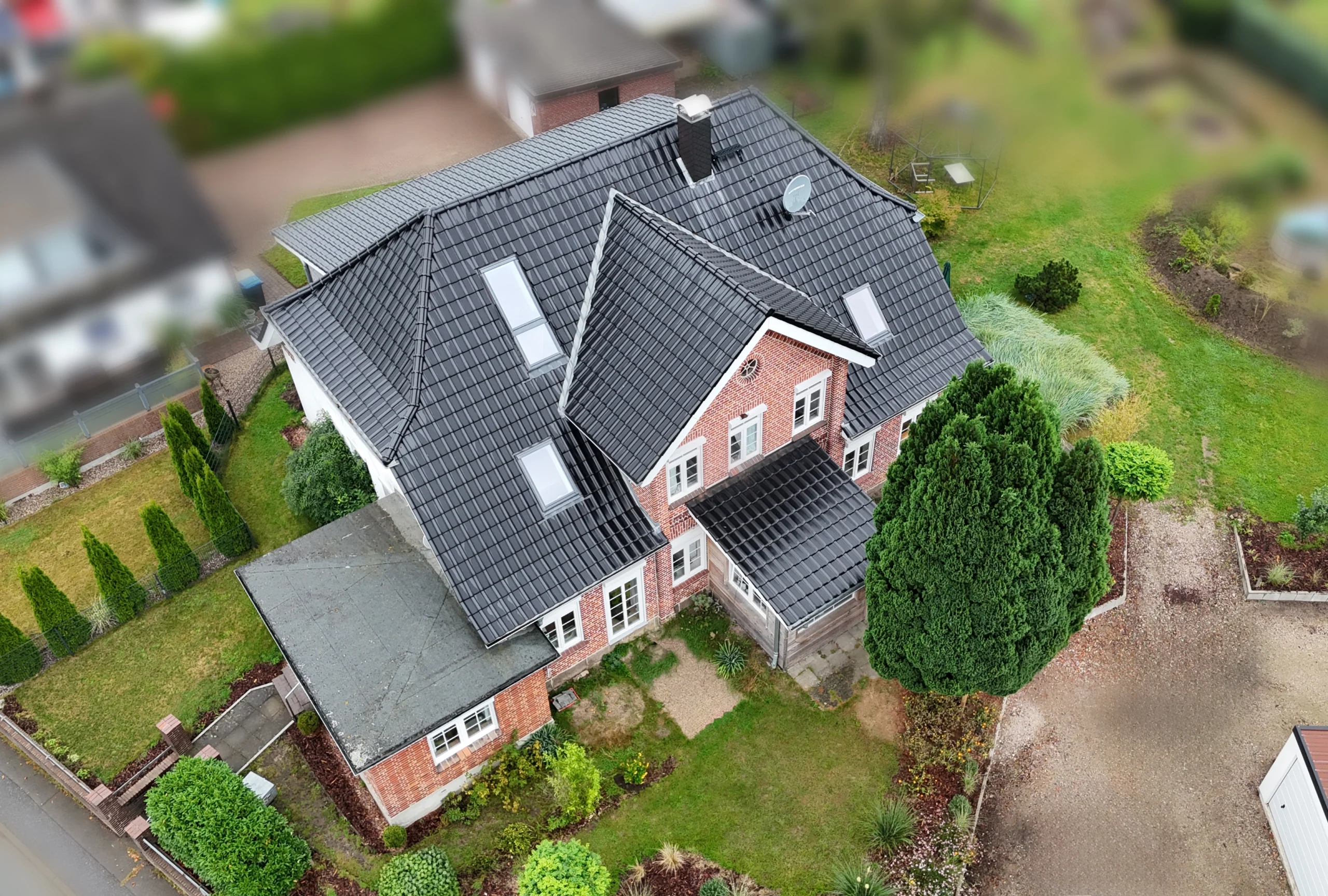 Luftaufnahme eines modernen Einfamilienhauses mit dunklem Ziegeldach, mehreren Dachfenstern und roter Klinkerfassade, umgeben von einem gepflegten Garten.