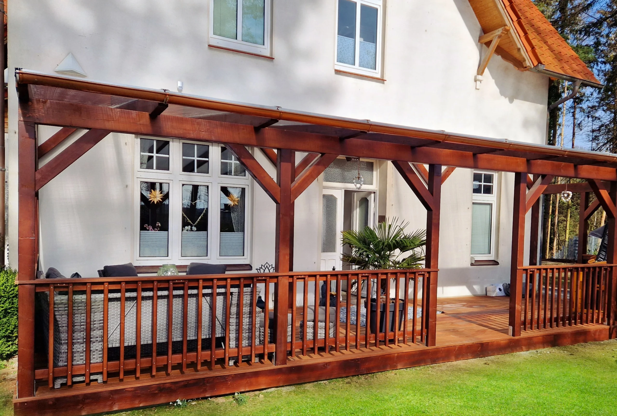 Eine maßgefertigte Holzpergola mit transparentem Dach und integrierter Holzterrasse, die an ein weißes Wohnhaus angebaut ist.