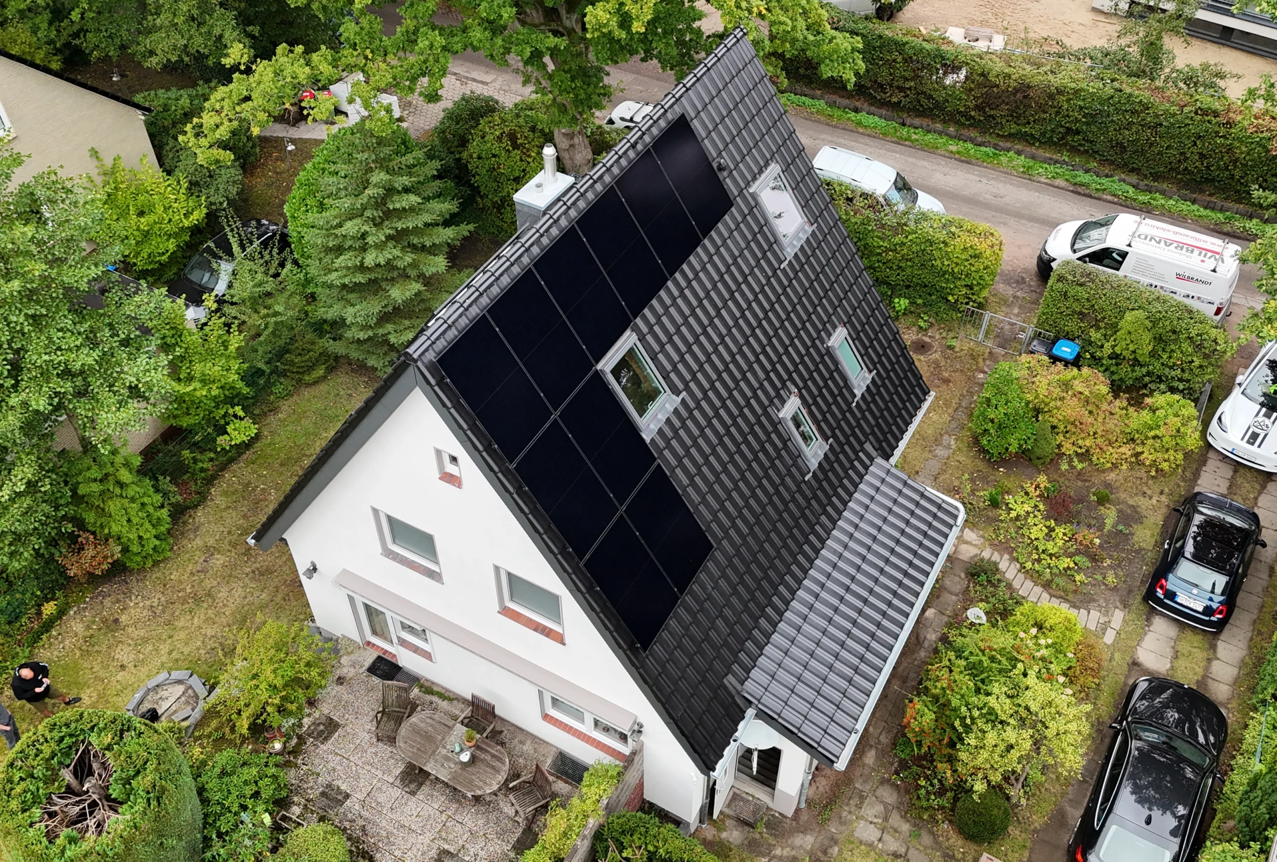 Luftaufnahme eines modernen Einfamilienhauses mit dunklem Ziegeldach, integrierten Solarmodulen und Dachfenstern, umgeben von einem grünen Garten.