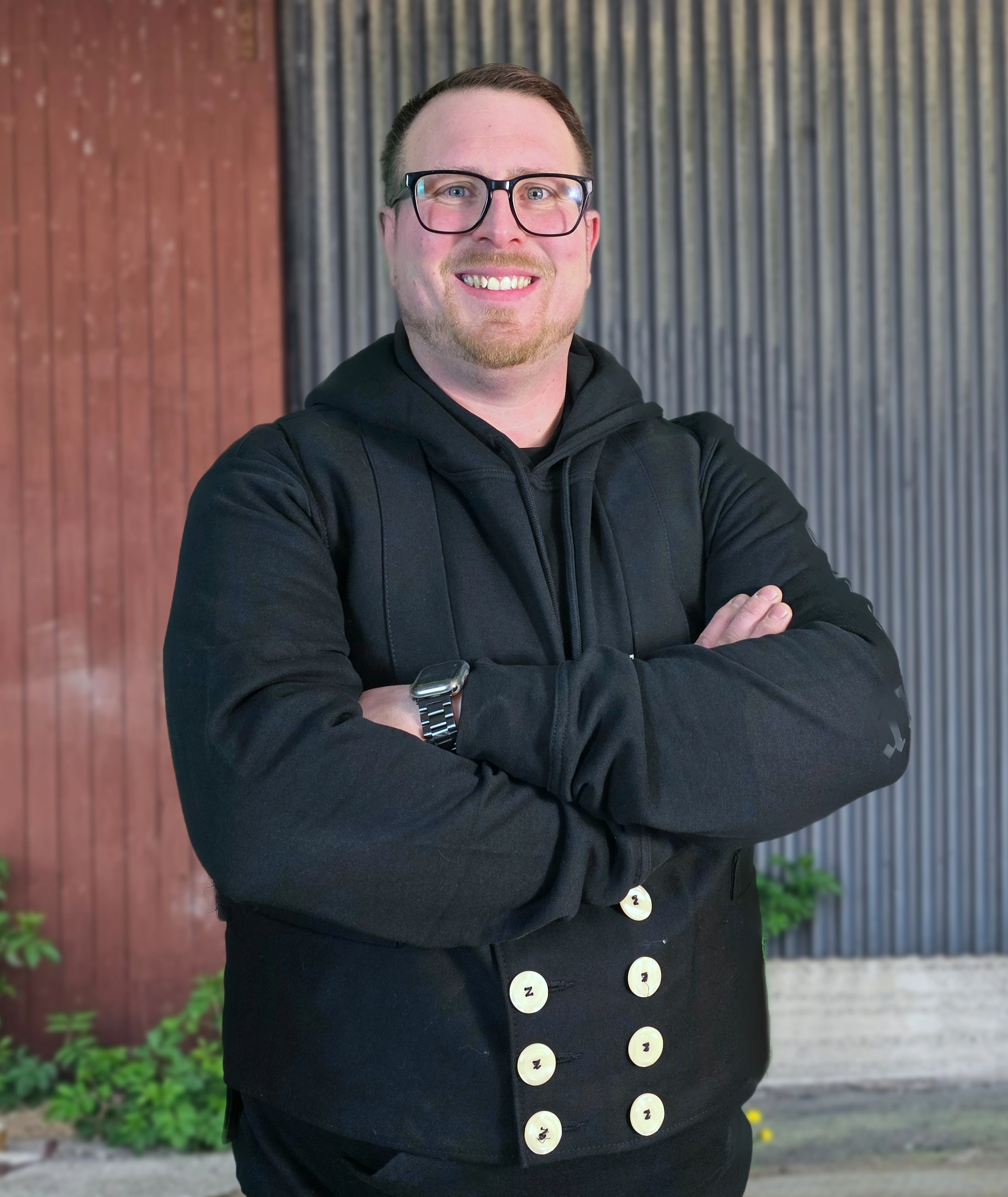 Sebastian Jordt, ein lächelnder Zimmermann mit Brille und traditioneller Zunftweste, steht mit verschränkten Armen.