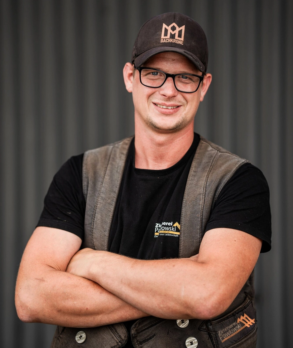Portrait von Sebastian Fey, einem Mitarbeiter der Zimmerei Pawlowski GmbH, der lächelnd mit verschränkten Armen in Arbeitskleidung posiert.