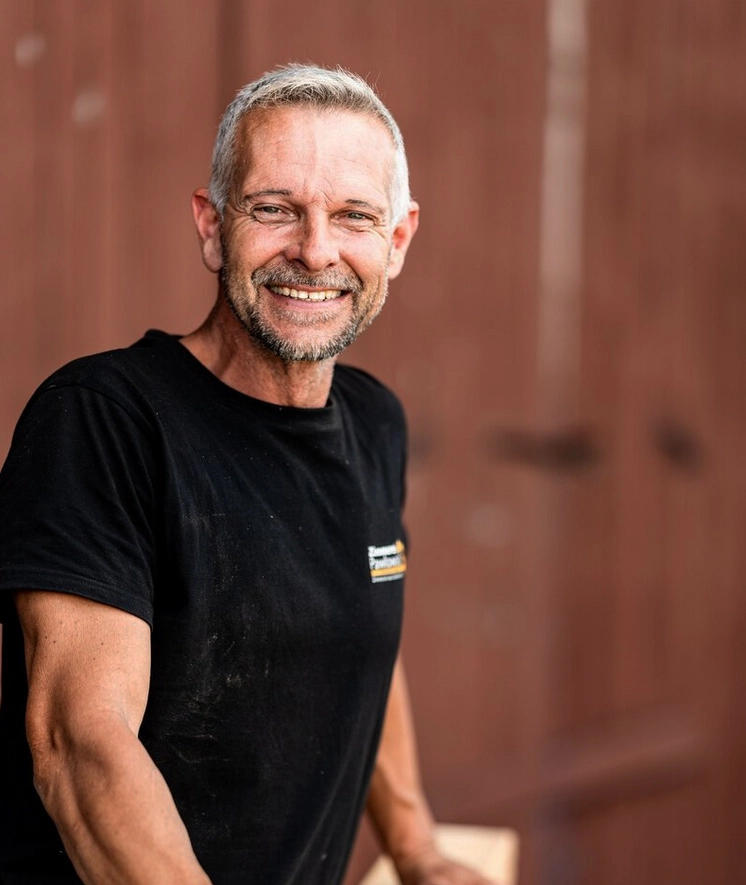 Porträt von Sven Schmädicke, einem lächelnden Mitarbeiter der Zimmerei Pawlowski GmbH, vor einem unscharfen, rötlich-braunen Hintergrund.