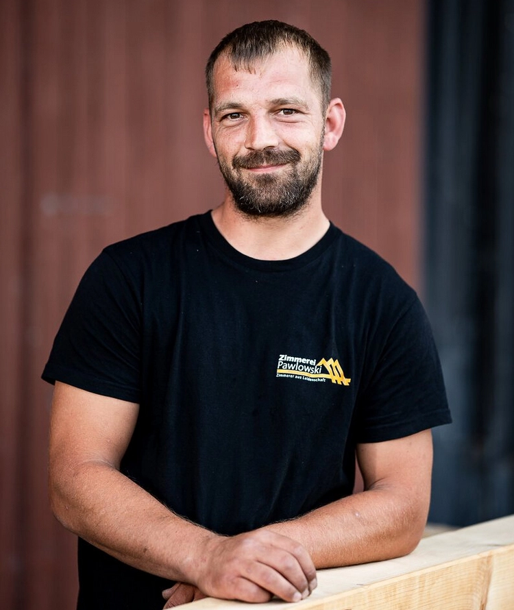 Portrait von Torben Westphal, einem Zimmermann der Zimmerei Pawlowski GmbH, der lächelnd auf einem Holzbalken lehnt.