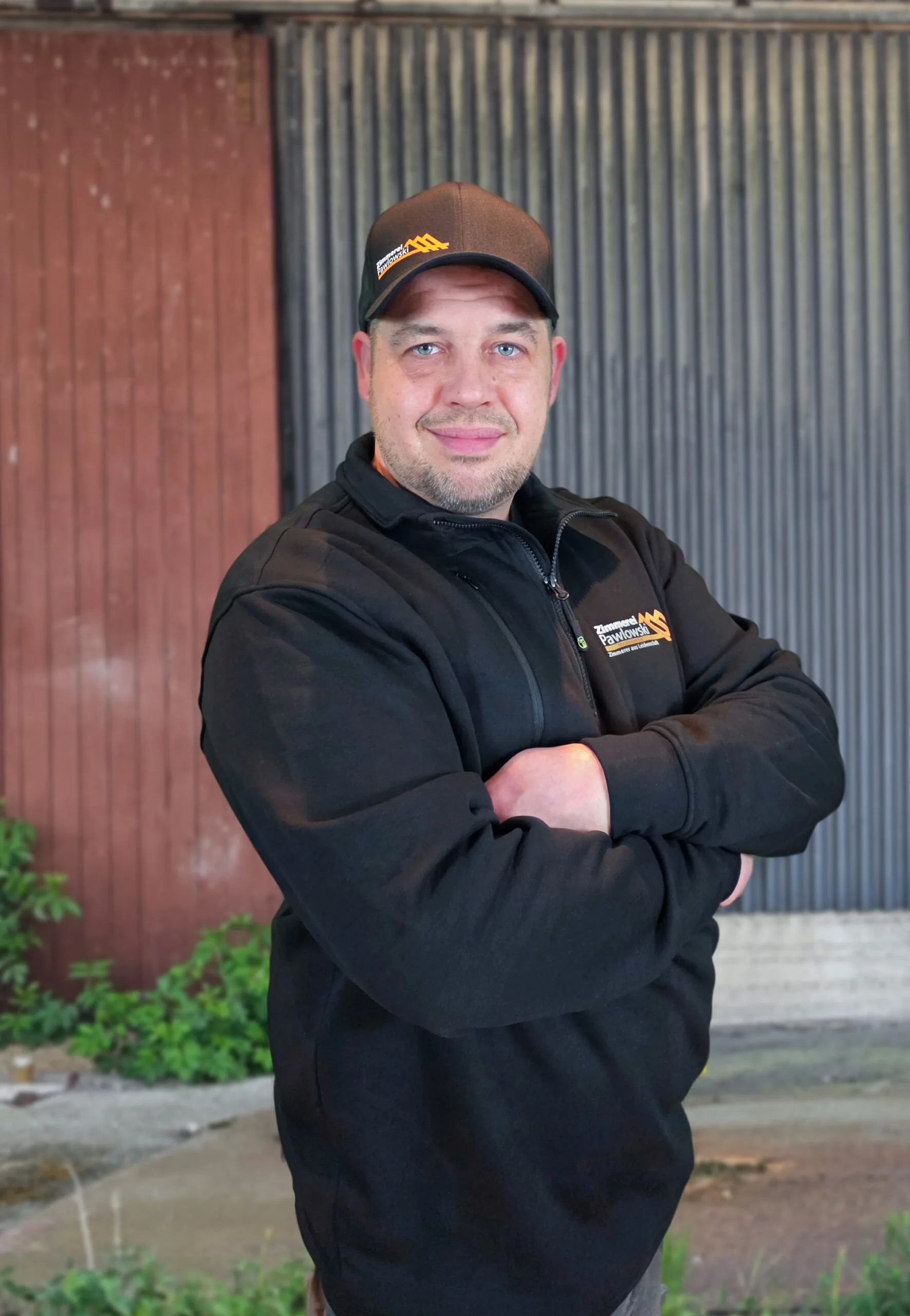 Ein erfahrener Zimmerer der Zimmerei Pawlowski GmbH posiert selbstbewusst in Arbeitskleidung mit verschränkten Armen vor einer Werkstattwand.